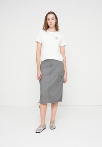 T-shirt blanc avec un petit logo, associé à une jupe grise longueur genou avec une poche latérale, complétés par des chaussures plates argentées.