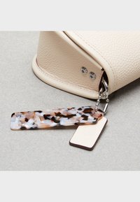 Kremowa torebka z teksturowanej skóry o zakrzywionym kształcie; zawiera wielokolorową, wzorzystą zawieszkę wykonaną z akrylu oraz metalowe ozdoby.