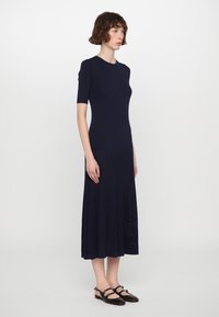 Abito a maglia blu navy con maniche corte, corpo aderente e gonna plissettata fluida. Abbinato a scarpe basse marroni scure con cinturini.