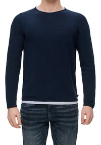 Mies, który nosi ciemnoniebieską fakturowaną koszulę z długim rękawem na białej podkoszulce, z ciemnoniebieskimi, lekko przetartymi dżinsami.