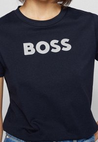 Námořnická modrá bavlněná tričko se velkým stříbrným glitrovým potiskem "BOSS" na přední straně. Klasický kulatý výstřih a krátké rukávy.