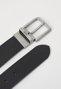 Curea din piele neagră cu cataramă din metal argintiu, branduită "BOSS." Catarama are o formă rectangulară și un finisaj mat.