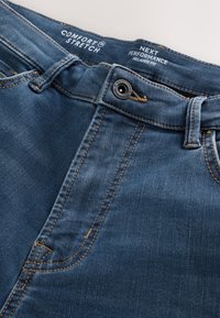 Jeans in denim blu con bottone, passanti per cintura e testo stampato all'interno della vita con la scritta "Comfort Stretch" e "Next Performance Relaxed Fit."