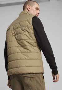Gilet imbottito khaki con design trapuntato e colletto alto; presenta un logo nero sulla parte posteriore, abbinato a una maglietta nera a maniche lunghe.
