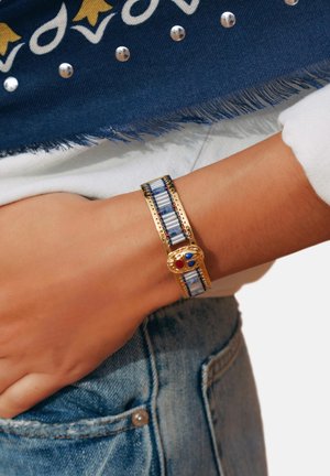 Bracelet en or avec un motif rayé bleu et blanc, comportant un fermoir décoratif avec des accents rouges et bleus. Porté au poignet.