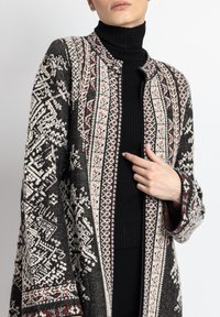 Sorte og hvide mønstrede cardigan med røde accenter, åben front, lange ærmer og en struktureret overflade, båret over en ribbet sort top.