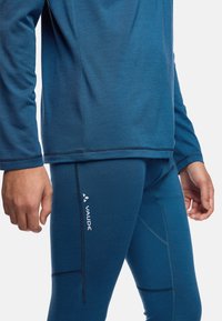 Camicia blu a maniche lunghe con texture liscia e orlo curvo, abbinata a leggings blu con dettagli di cucitura e logo "Vaude".