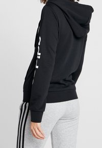 Femme portant un sweat à capuche noir avec du texte blanc sur la manche et un pantalon de survêtement gris clair avec des bandes noires sur les côtés, vue de dos.