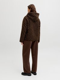 Person som står, iført en brun mønstret hettejakke, løstsittende brune bukser og svarte sko mot en ensfarget lys bakgrunn.