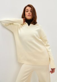 Pulover tricotat de culoare crem, cu gât în V și manșete cu model din răsucire. Asortat cu o pereche de pantaloni corespunzători. Textură fină, croi lejer, detalii minime.