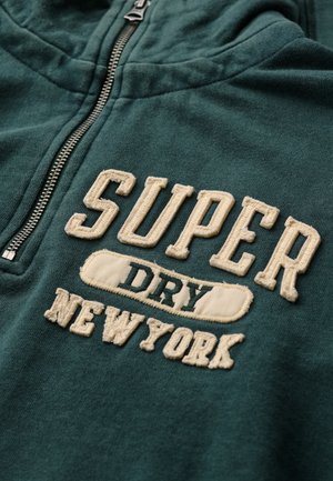 Sudadera verde con cremallera frontal. Cuenta con texto bordado en blanco que dice "SUPER DRY NEW YORK." Tejido suave y diseño con capucha.