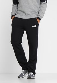 Svarta svettbyxor med lös passform, med en vit Puma-logga. Tyget är mjukt med en slät yta, kombinerat med en grå sweatshirt.