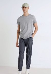T-shirt gris à manches courtes, pantalon élastique gris foncé, et casquette vert clair. Le modèle se tient devant un fond uni.