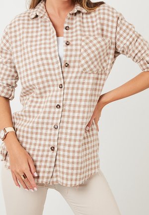 Femme portant une chemise à carreaux beige et blanche à boutons avec poche poitrine, un sous-t-shirt blanc, un pantalon beige, une montre-bracelet et une bague, main sur la hanche.