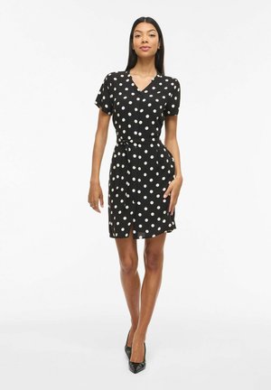 Kobieta stoi na białym tle, ubrana w czarną sukienkę z krótkim rękawem w białe grochy, z paskiem wiązanym w talii oraz czarne szpilki z ostrymi noskami.