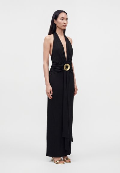 Woman standing in a black halter-neck gown with a deep V-neck and gold circular buckle at the waist, wearing gold open-toe heels.