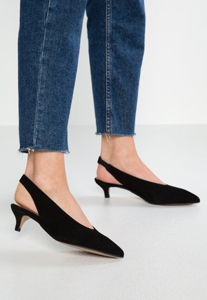 Nohy v černých semišových botách na podpatku s otevřenou patou, spojené s zkrácenými modrými džínami s třepenými okraji.