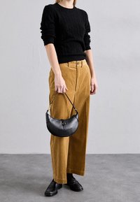 Schwarzer Kabelstrickpullover, oversize Schnitt. Helle Cord-Hosen mit weitem Bein und vorderen Taschen, kombiniert mit einer kleinen schwarzen Ledertasche. Schwarze Loafers.