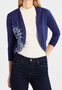 Laivastonsininen neuletakki, jossa on avoin etu, vasemmalla puolella monimutkainen valkoinen kukkaommelu ja kolmen neljänneksen hihat, yhdistetty kermaiseen toppiin.