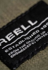 Etichetta in tessuto nera con testo bianco che legge "REELL FONDATO NEL 1993" e "DA ZERO PROGETTATO DA UNITED" su uno sfondo verde oliva.