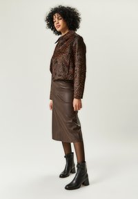 Kobieta z kręconymi włosami, ubrana w brązową kurtkę w panterkę, skórzaną spódnicę do kolan w kolorze brązowym, czarne przezroczyste rajstopy i czarne botki za kostkę.