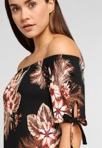 Top floral de hombros caídos con tejido negro, presenta grandes flores blancas y coral, mangas cortas y detalles de lazo en las mangas.