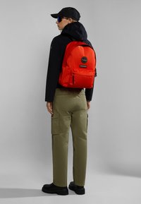 Zaino rosso con tasca frontale, spalline regolabili e marchio, indossato sopra una giacca nera e pantaloni cargo verde oliva, con una vestibilità dritta.
