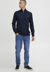 Pull-over marine à manches longues avec une fermeture éclair quart, tissu texturé et bords côtelés, associé à un jean léger à jambe droite bleue et des baskets noires.