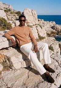 Homme portant des lunettes de soleil, une veste beige zippée, un pantalon blanc et des mocassins marron, assis sur une côte rocheuse avec la mer bleue en arrière-plan.