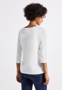 Witte, relaxte top met driekwartmouwen en een wijd decolleté. Gladde katoenstof met minimale details. Draag het met donkere jeans.