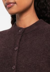 Femme portant un cardigan marron foncé à boutons, duveteux, avec un col rond, montrant le bas du visage et le cou.