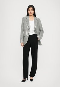 Donna che indossa un blazer grigio con paillettes sopra una maglietta bianca, pantaloni neri a gamba larga e tacchi a punta neri, in piedi con una mano in tasca.