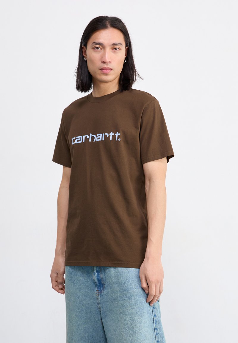 Hnedé bavlnené tričko s bielym logom "carhartt." Krátke rukávy a klasický strih s okrúhlym výstrihom. Voľný strih s jemnou textúrou.