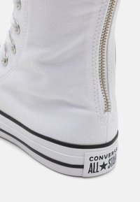 Gros plan d’une basket blanche montante Converse All Star montrant la texture du tissu, les œillets métalliques, la fermeture éclair latérale et la semelle en caoutchouc marquée.