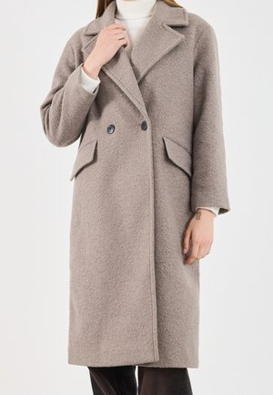 Femme portant un long manteau en laine taupe avec de larges revers, double boutonnage et poches à rabat sur un pull col roulé blanc.