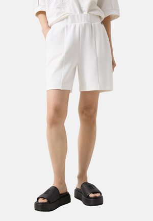 Person trägt weiße, hoch taillierte, maßgeschneiderte Shorts mit vorderen Falten und schwarze Plateau-Sandalen zum Hereinschlüpfen, steht mit den Händen in den Taschen.