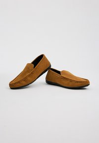 Mocasines de ante marrón con suelas de goma negras planas, que presentan un detalle de costura en la parte delantera y una textura suave, diseñados para un uso casual.