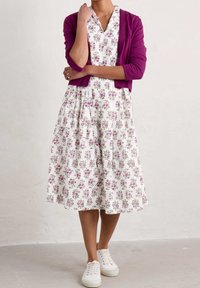 Vit blommig klänning med lila detaljer, med korta ärmar, V-ringning och en utsvängd kjol, i kombination med en lila cardigan och vita sneakers.