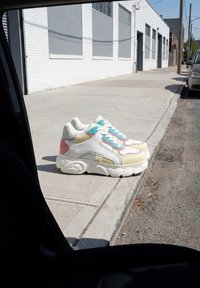 Par de zapatillas coloridas colocadas en el borde de una acera junto a una calle con coches aparcados y edificios de ladrillo blanco al fondo.