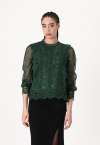 Blusa verde in pizzo con bordi scallop, maniche lunghe trasparenti e intricati motivi floreali. Completata con un collo e polsini a coste.
