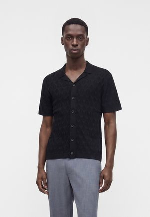 Homme portant une chemise noire à manches courtes boutonnée avec un motif texturé subtil et un pantalon gris clair, debout devant un fond uni.