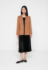 Blazer marrone sartoriale con rever a notched, gonna nera plissettata, ballerine nere e borsa rosa chiaro. Linee semplici e pulite e texture morbide.