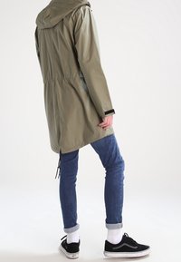 Personne portant une parka olive, un jean bleu retroussé, des chaussettes blanches et des baskets noires, se tenant contre un fond blanc uni.