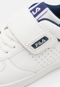 Sneaker in pelle bianca con cinturino in velcro, dettagli in blu navy, forature laterali e etichetta sulla linguetta in blu navy e bianco.