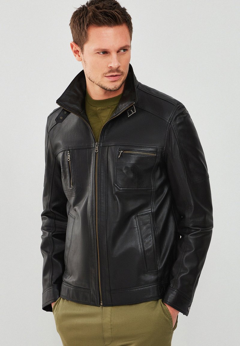 Giubbotto in pelle nera con colletto alto, tasche con zip e dettagli cuciti. Presenta una texture liscia e un design su misura.
