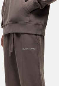 Brown sweatshirt and matching sweatpants with a smooth texture. Sweatpants feature an embroidered logo on the pocket. Drawstring detail visible.