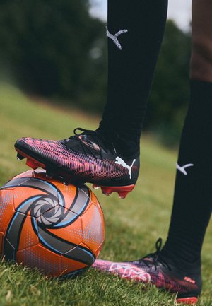 Mustad jalgpalliketsad esitlevad dünaamilist roosa ja lilla mustrit, mis on paigutatud oranži ja musta mustriga jalgpallile haljasalal.