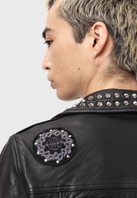 Jachetă neagră de piele cu ținte argintii pe umăr, având o insignă rotundă brodată cu un design de lanț și numele mărcii.