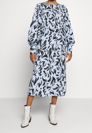 Mujer con un vestido midi azul claro con estampados abstractos en negro y mangas largas abullonadas, combinado con botines blancos hasta el tobillo.
