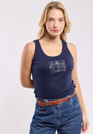 Femme blonde portant un débardeur bleu marine à motif carré abstrait blanc, rentré dans un jean taille haute bleu avec une ceinture marron.